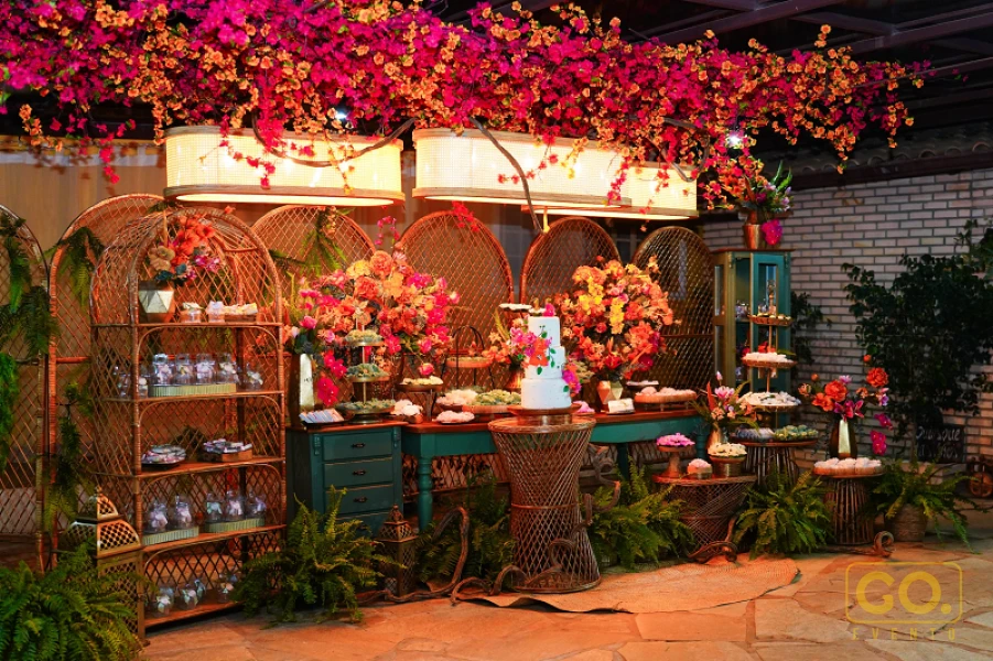 Mesa de doces decorada com bolo branco de andares, arranjos de flores e móveis de vime sob flores cor-de-rosa suspensas.