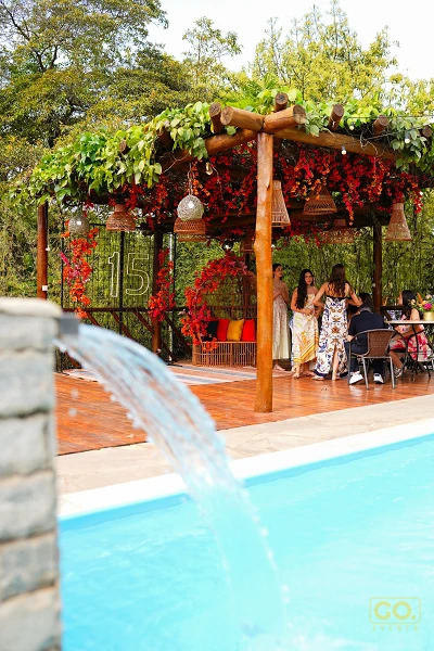Pérgola de madeira decorada com flores vermelhas ao lado de uma piscina com cascata, com convidados conversando em mesas ao ar livre.