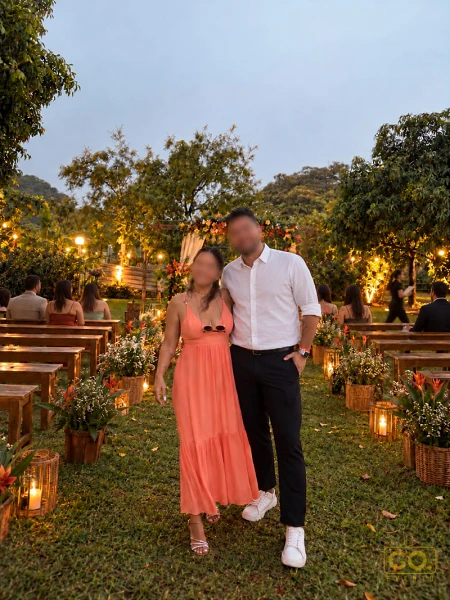 Casal caminha pelo corredor entre bancos de madeira de uma cerimônia ao ar livre, com luminárias e flores no gramado ao entardecer.