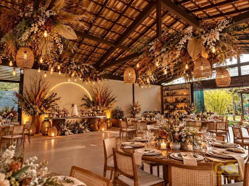 Salão coberto com telhado de madeira, pendentes de vime e flores suspensas, mesas redondas postas e mesa de bolo ao fundo.