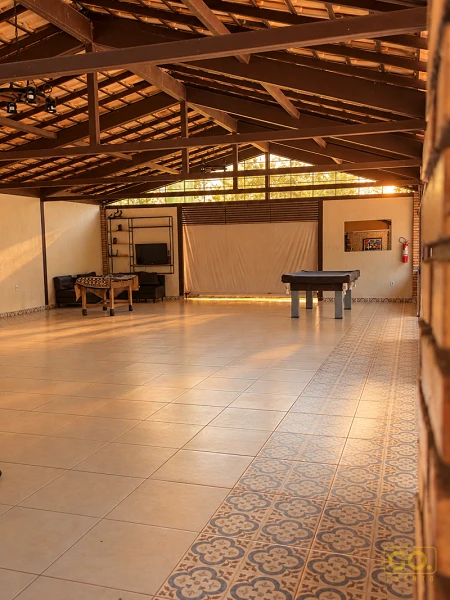 Salão coberto com piso de ladrilho decorado, mesa de sinuca e pebolim, teto de madeira e janelas altas ao fundo.