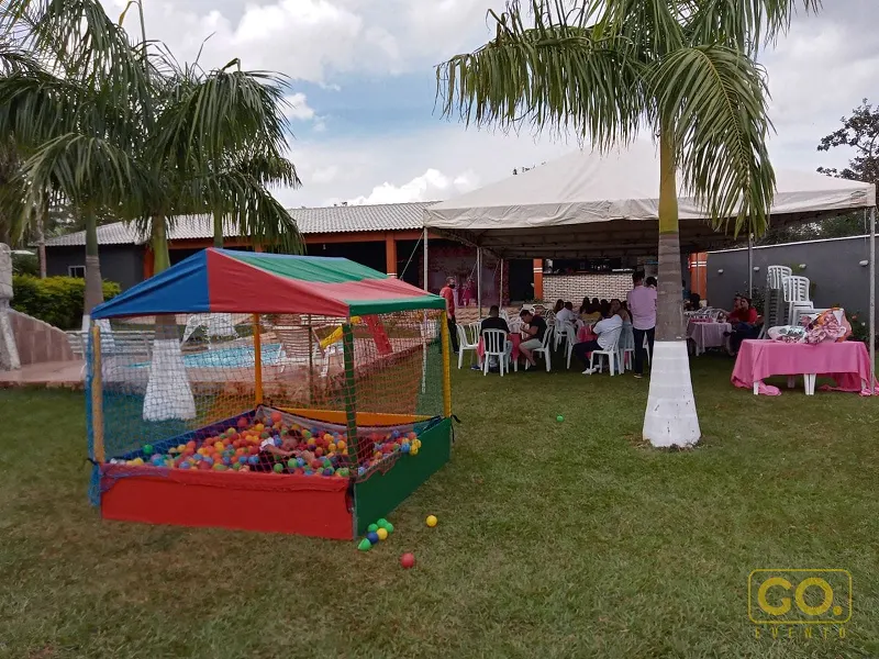 Tenda branca sobre cadeiras e mesas com toalha rosa, ao lado de piscina de bolinhas coloridas no gramado com palmeiras.