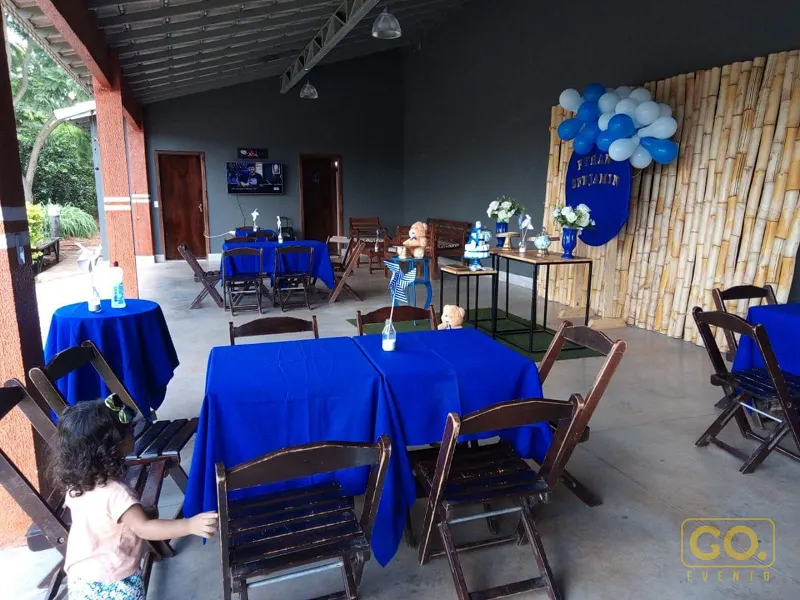 Área coberta decorada com toalhas azuis, cadeiras de madeira e arco de balões azuis e brancos ao lado de painel de bambu.