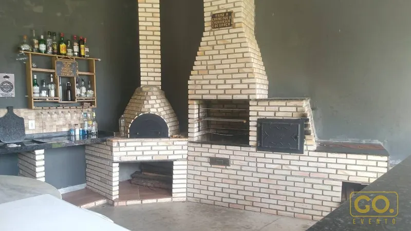 Churrasqueira e forno de pizza de tijolos aparentes em área gourmet coberta, com balcão de granito e bar com garrafas ao fundo.