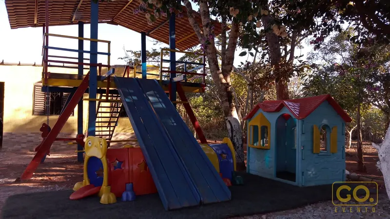 Parquinho ao ar livre com escorregador azul, plataforma sob telhado listrado e casinha de plástico azul entre árvores.