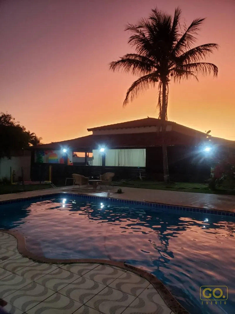 Piscina ao entardecer com borda ladrilhada, palmeira alta e casa térrea com varanda iluminada ao fundo.