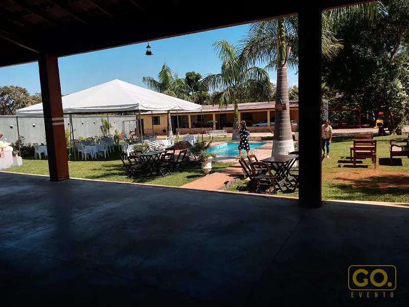 Vista de área coberta para o gramado, com piscina, mesas de madeira, cadeiras brancas e uma lona branca montada ao fundo.