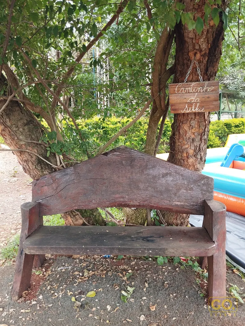 Banco rústico de madeira sob árvores, com placa para selfie pendurada no tronco e um brinquedo inflável ao lado.
