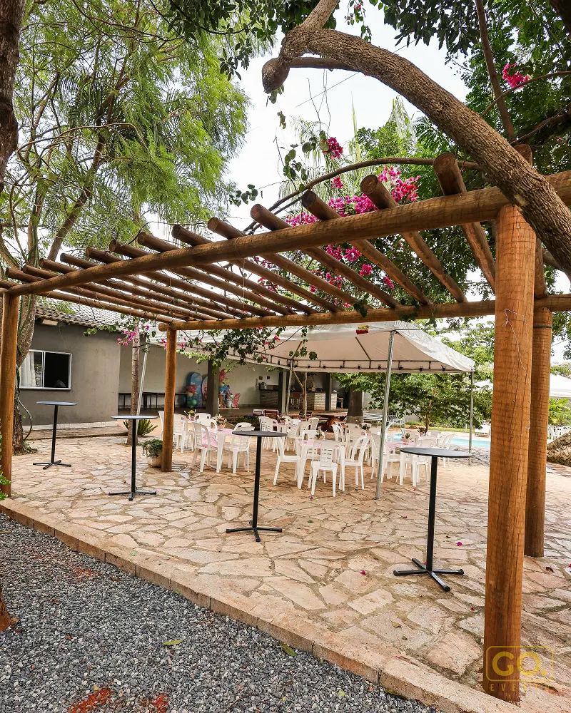 Pérgola de madeira sobre piso de pedra, com mesas altas, cadeiras brancas, toldo e piscina ao fundo sob árvores floridas.