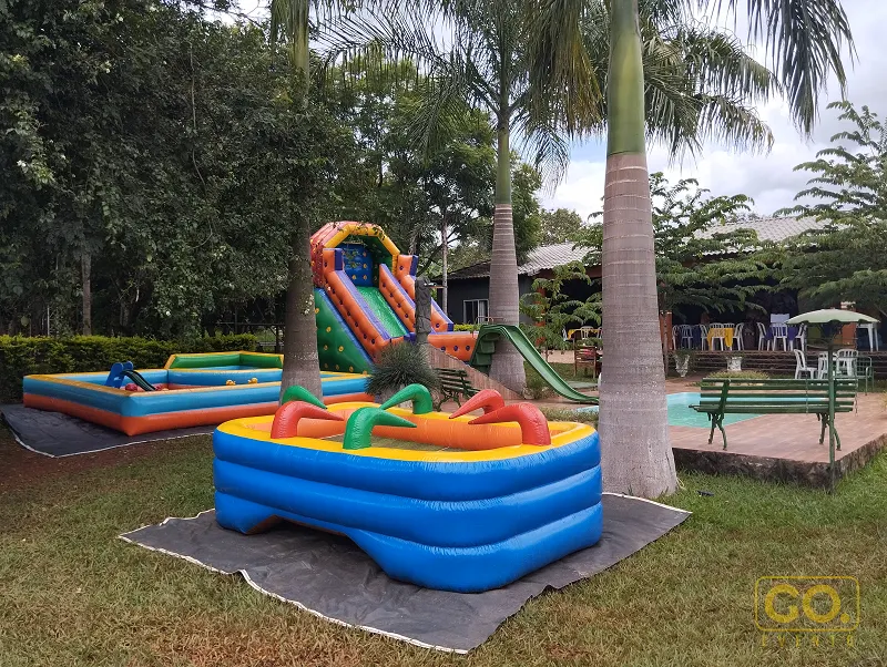 Brinquedos infláveis coloridos, incluindo um escorregador e uma piscinha inflável, montados sobre a grama perto de uma piscina.
