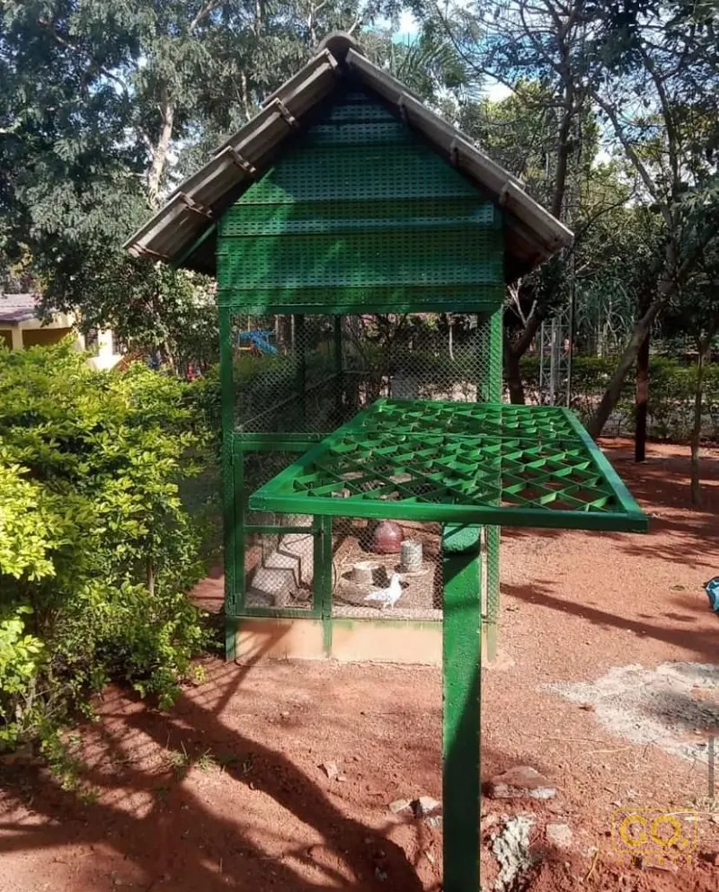 Viveiro metálico verde com telhado e tela de arame, com uma ave branca no chão de terra avermelhada entre árvores.