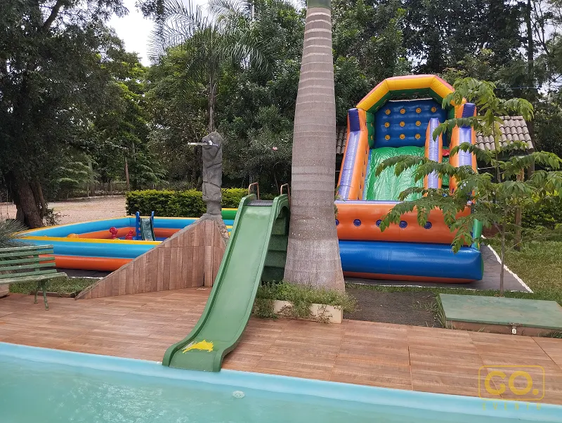 Piscina ao ar livre com deck de madeira, piscina infantil inflável colorida e escorregador inflável ao lado de palmeira.