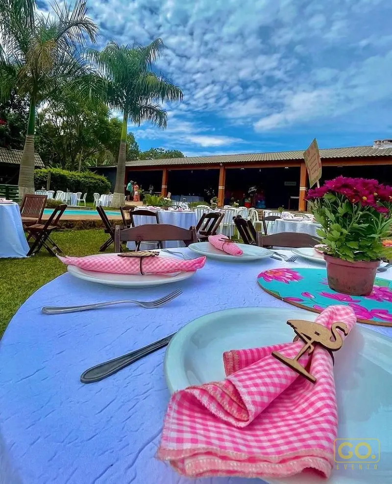 Mesa redonda posta com toalha azul, guardanapos xadrez rosa e enfeites de flamingo de madeira, com piscina e palmeiras ao fundo.