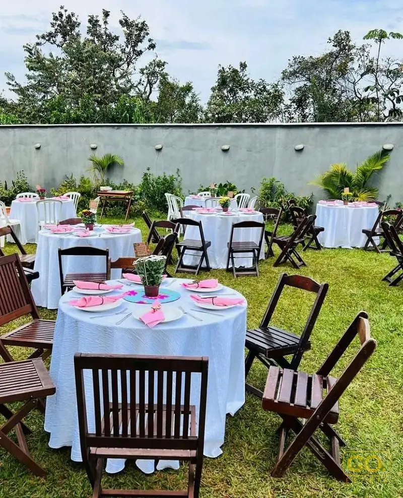 Mesas redondas com toalhas brancas e guardanapos rosa montadas em um jardim gramado, com árvores e muro cinza ao fundo.