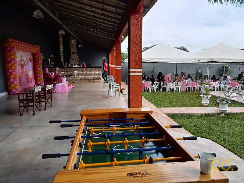 Área coberta com arco de balões rosa e dourados, bar de tijolinhos e mesa de pebolim, com convidados ao lado do gramado.