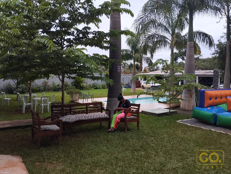 Gramado com palmeiras e bancos de madeira ao lado de uma piscina e de um brinquedo inflável colorido ao fundo.
