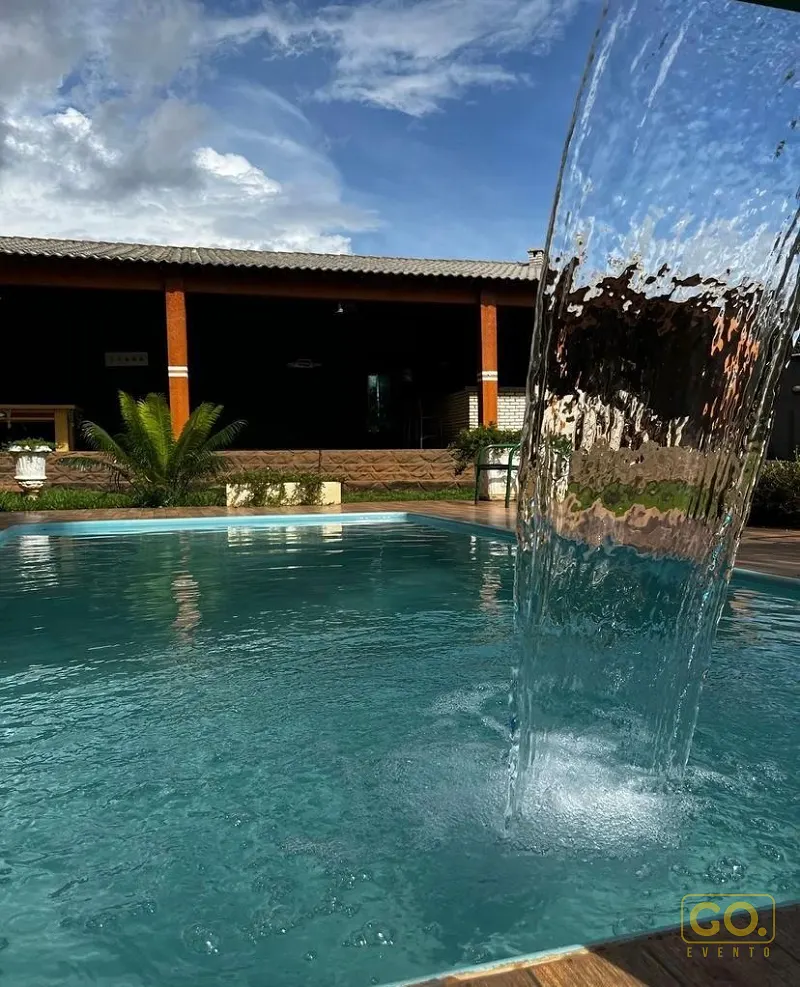 Piscina de água azul com cascata de parede em primeiro plano e casa rústica de colunas de madeira ao fundo, sob céu azul.