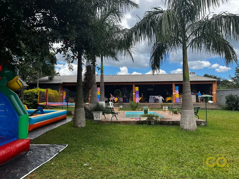 Gramado com piscina pequena, palmeiras e casa térrea de colunas laranja decorada com balões para festa infantil.