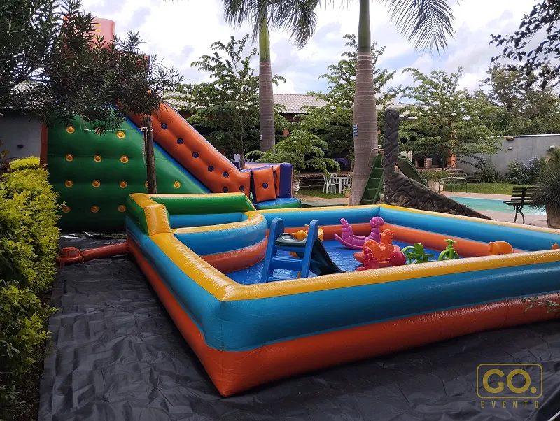 Área infantil ao ar livre com piscina inflável colorida, escorregador e parede de escalada infláveis sob palmeiras.