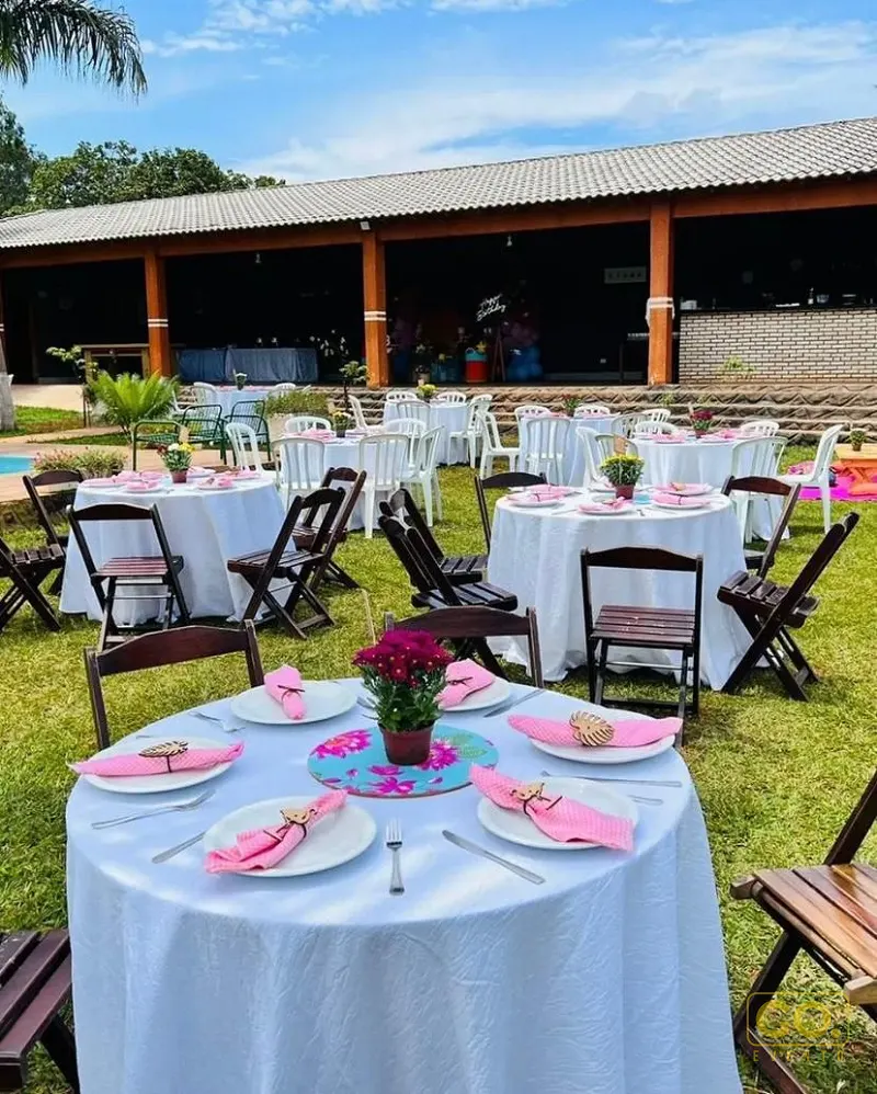 Mesas redondas com toalhas brancas, guardanapos rosa e flores sobre o gramado, com salão coberto de madeira ao fundo.