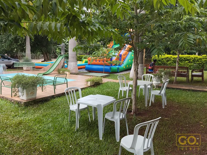 Área verde com gramado, mesas e cadeiras brancas de plástico, piscina ao fundo e brinquedo inflável colorido entre as árvores.