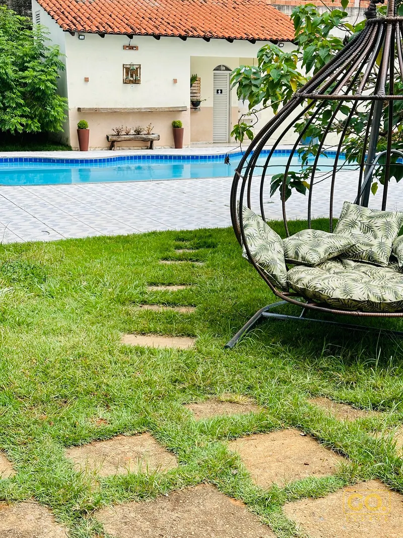 Piscina azulejada ao lado de gramado com piso de pedra e uma poltrona suspensa com almofadas de folhagens verdes.