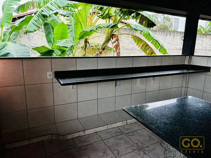 Bancada de granito preto e azulejos claros em área gourmet coberta, com vista para bananeiras pela janela ao fundo.