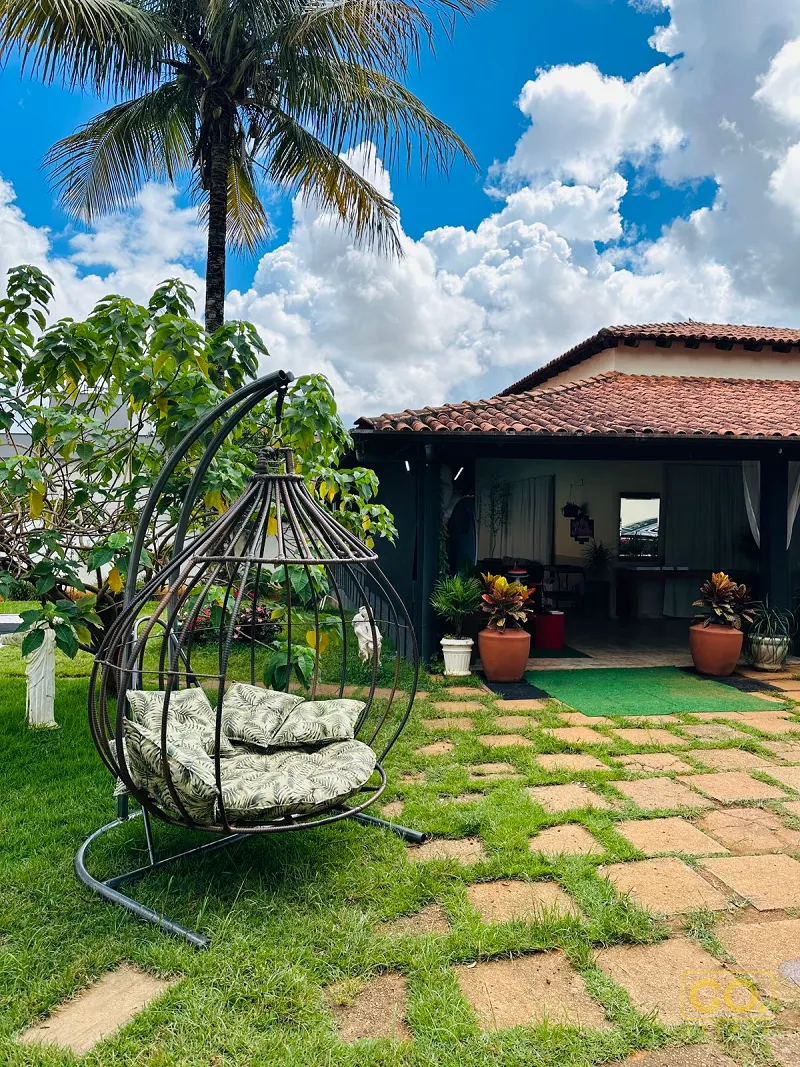 Jardim com gramado, piso de pedra e cadeira suspensa com almofadas, ao lado de uma casa com varanda e telhado de barro.