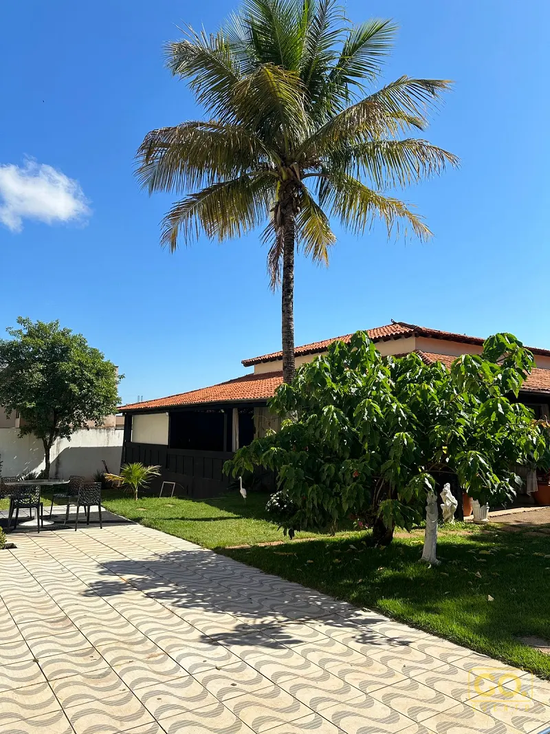 Gramado e calçada de piso ondulado ao lado de uma casa de telhado de barro, com palmeira alta e mesa de jardim à esquerda.