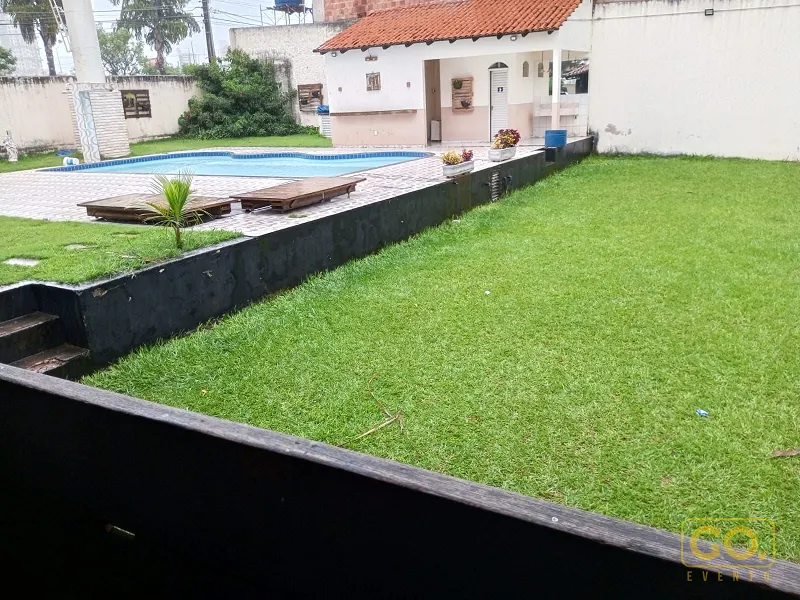 Gramado verde extenso ao lado de uma piscina com borda de azulejos e espreguiçadeiras de madeira, em dia nublado.