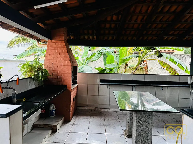 Área gourmet coberta com churrasqueira de tijolo aparente, bancada de granito, pia e mesa de vidro, com bananeiras ao fundo.
