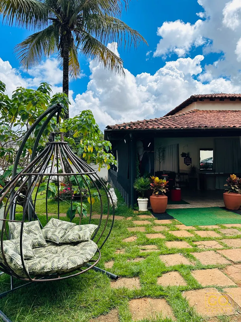 Jardim com piso de pedra entre a grama, cadeira suspensa de ferro com almofadas e casa de telhado cerâmico sob palmeira.