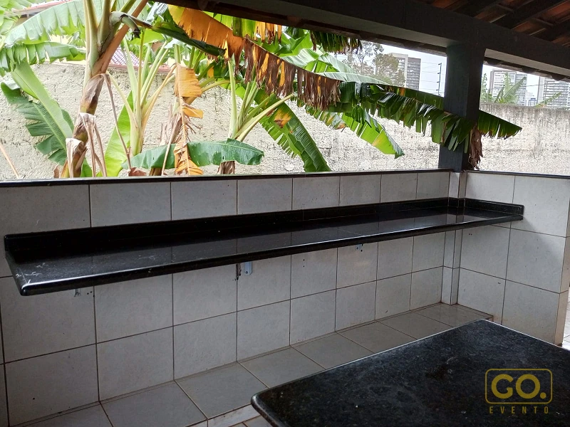 Bancada de granito preto em área coberta com azulejos brancos, e bananeiras ao fundo através da abertura lateral.