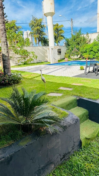 Jardim com gramado, degraus de grama sintética, piscina de borda azul ao fundo e caixa d'água branca em área externa ensolarada.